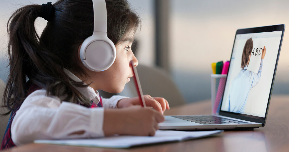 A young girl wearing headphones sits at a desk. She looks at a laptop - on its screen is an image of a teacher writing A, B, C - and then takes notes on a pad of paper.